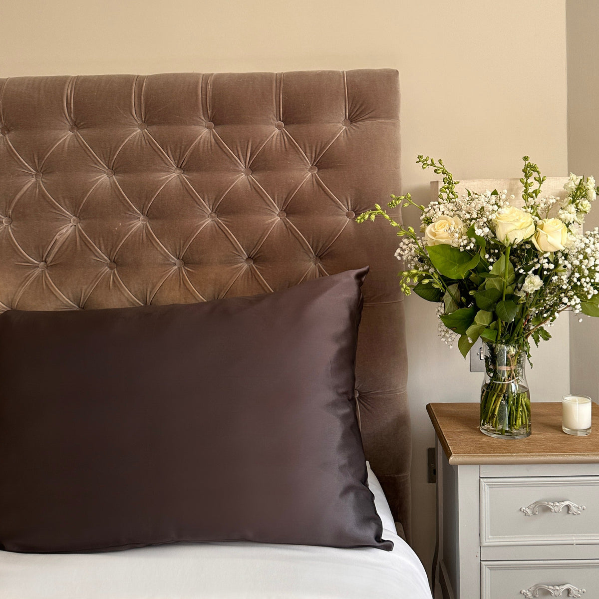 Chocolate brown mulberry silk pillowcase on a light velvet headboard beside a bedside table with flowers
