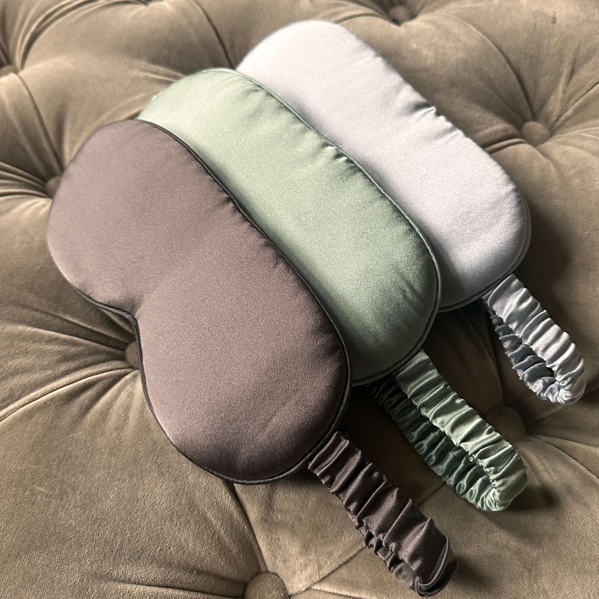 Pistachio, pearl white and oyster silk eye masks lined up on a velvet sofa